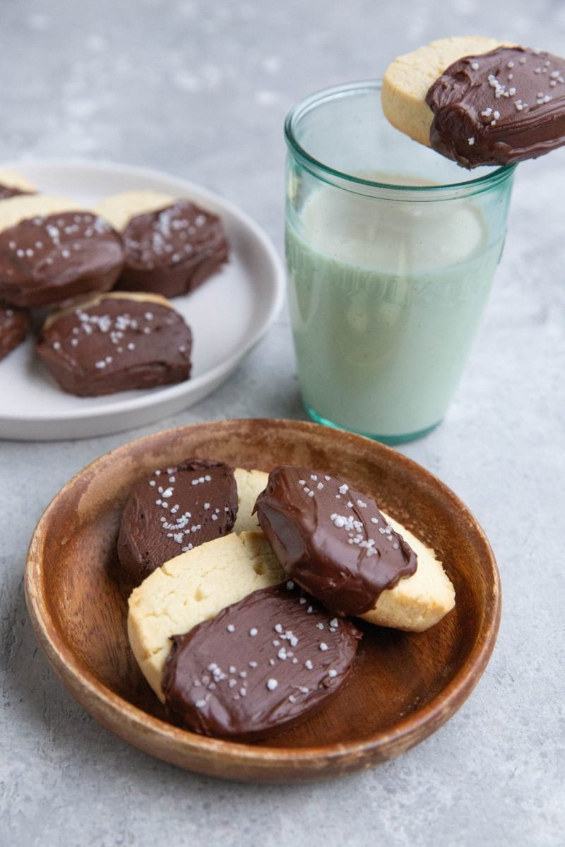 Two plates of chocolate dipped shortbread cookies with a glass of milk with a cookie on top.