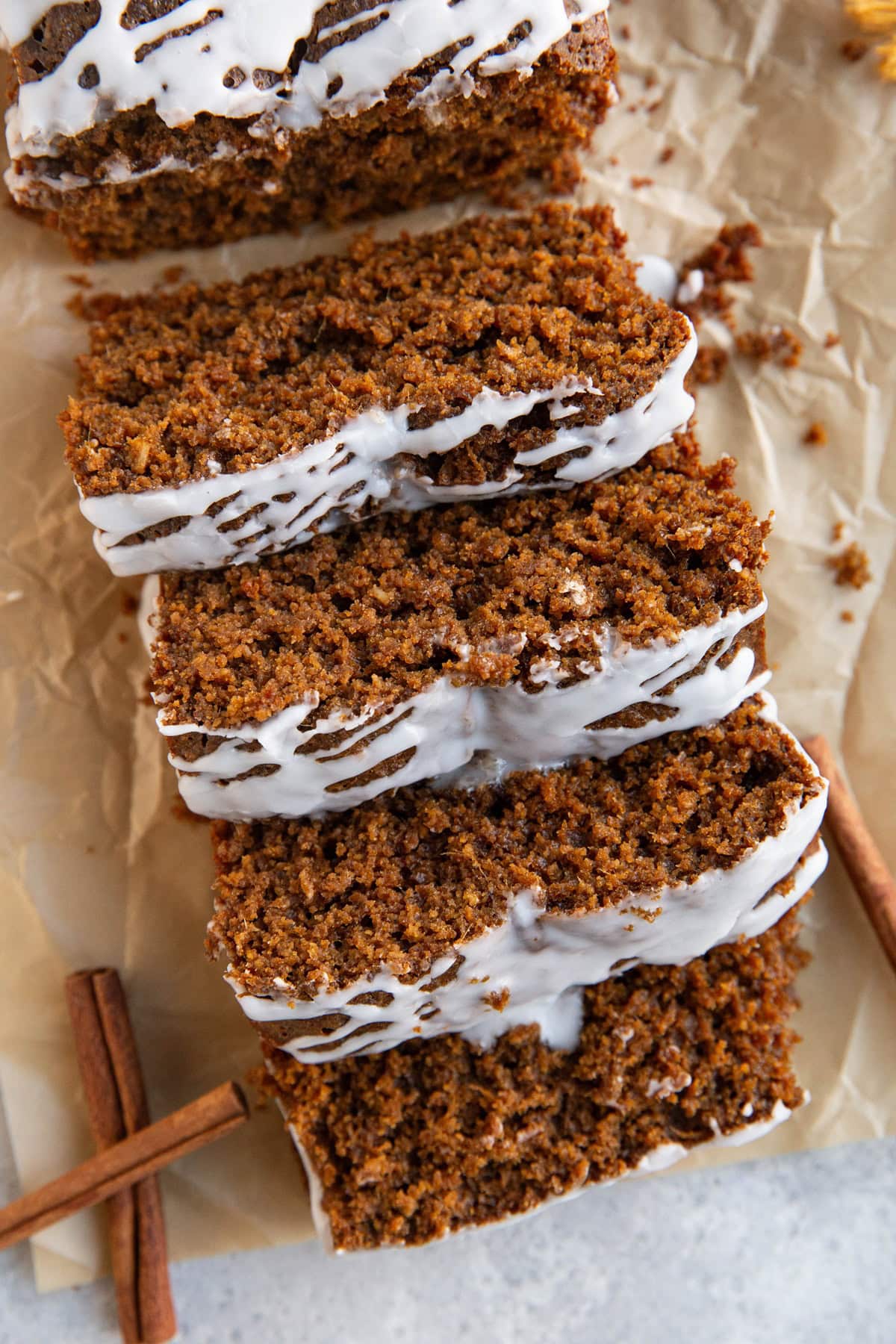 Gingerbread loaf seen from the top down view cut into slices with a glaze drizzled on top. A few cinnamon sticks to the side.