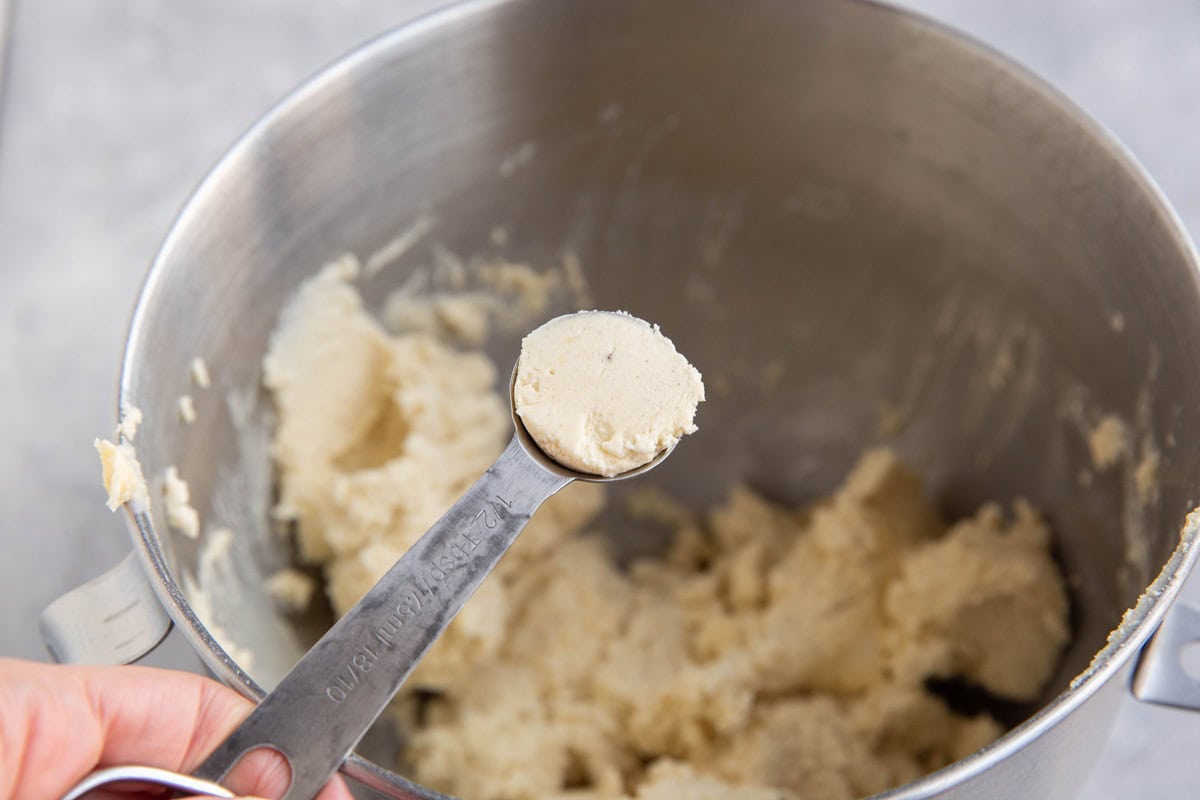 Bowl of a stand mixer full of cookie dough with a 1/2 Tbsp measuring spoon measuring out some dough.