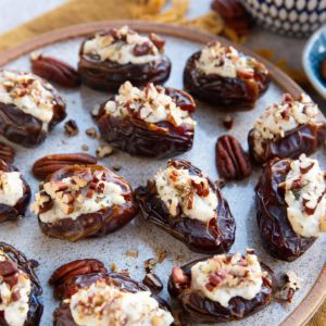Ricotta Stuffed Dates sprinkled with pecans sitting on a plate with a bowl of fresh dates in the background and a golden napkin, ready to serve!