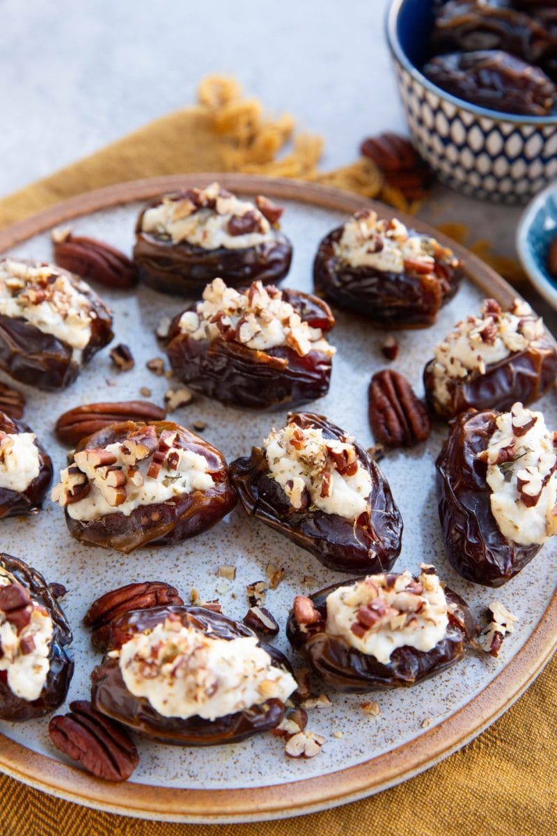 Ricotta Stuffed Dates sprinkled with pecans sitting on a plate with a bowl of fresh dates in the background and a golden napkin, ready to serve!