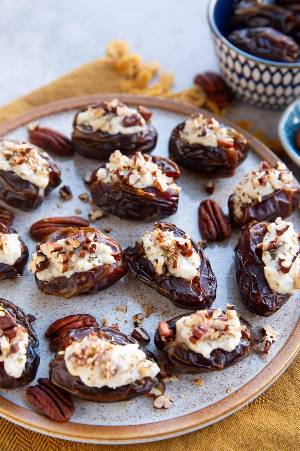 Ricotta Stuffed Dates sprinkled with pecans sitting on a plate with a bowl of fresh dates in the background and a golden napkin, ready to serve!