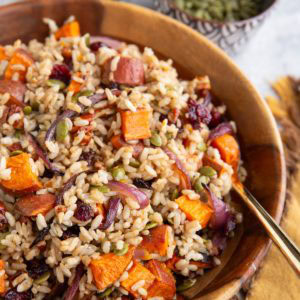 Large wooden bowl full of brown rice salad with roasted sweet potatoes, onions, dried cranberries, pecans. A bowl of pecans in the background and a bowl of pumpkin seeds. A golden napkin to the side, and a golden spoon sits inside the bowl, ready to serve.