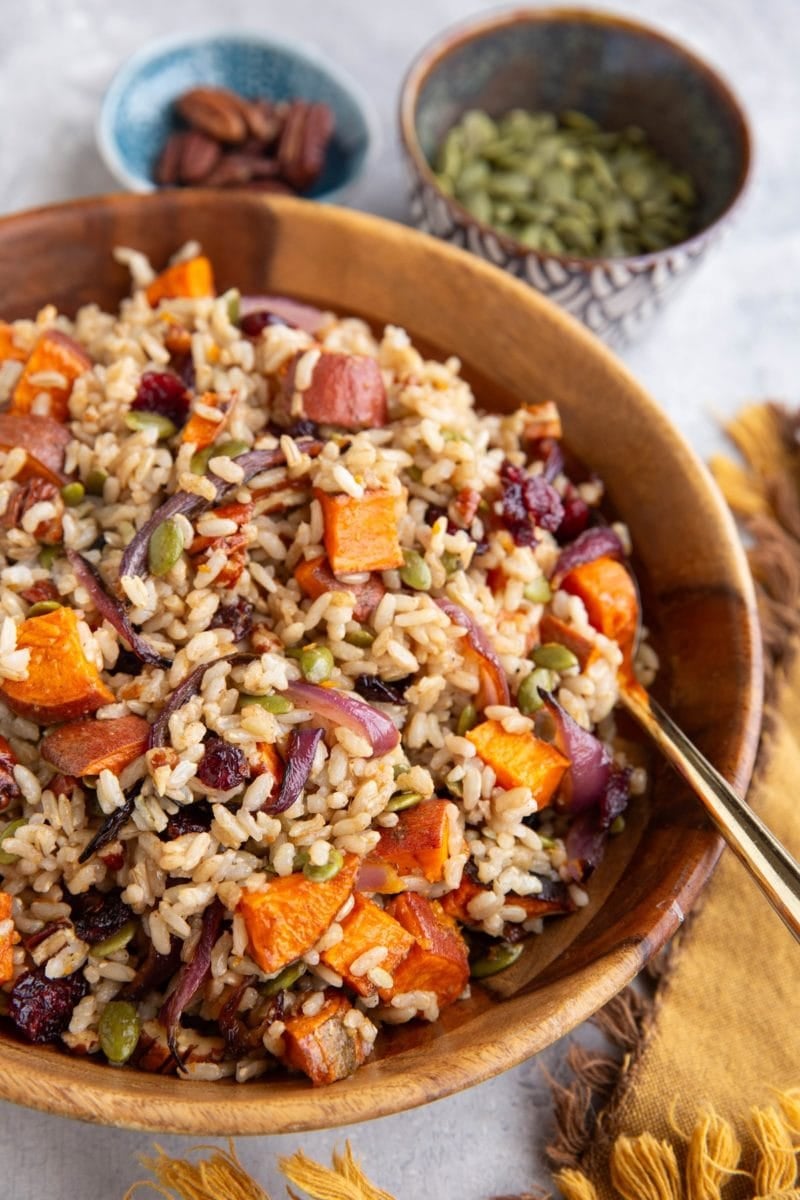 Large wooden bowl full of brown rice salad with roasted sweet potatoes, onions, dried cranberries, pecans. A bowl of pecans in the background and a bowl of pumpkin seeds. A golden napkin to the side, and a golden spoon sits inside the bowl, ready to serve.