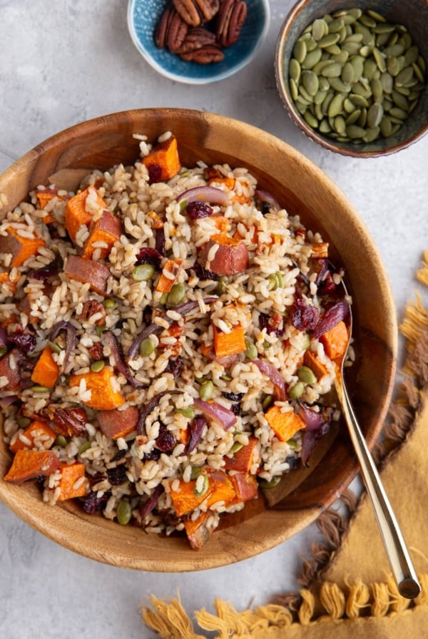 Brown rice salad in a wooden bowl with pumpkin seeds and pecans to the side in small bowls. Ready to serve.