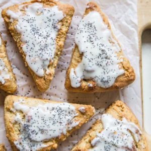 6 scones on a wooden cutting board sitting on top of parchment paper with a glaze and poppy seeds sprinkled on top.