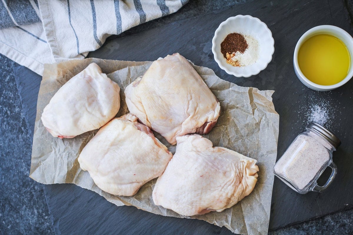 Raw chicken thighs on parchment paper next to a salt shaker, a small bowl of oil and a small bowl of seasonings.