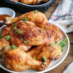 Plate of baked chicken thighs and drumsticks seasoned with a dry rub and sprinkled with chopped parsley. the rest of the baking dish of chicken in the background and a burlap napkin to the side.
