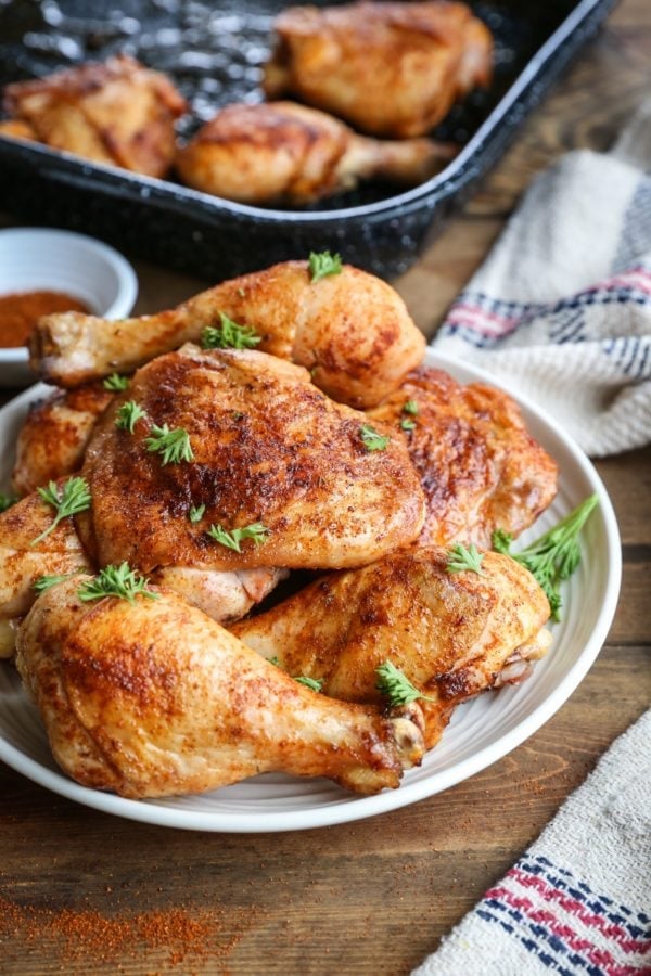 Plate of baked chicken thighs and drumsticks seasoned with a dry rub and sprinkled with chopped parsley. the rest of the baking dish of chicken in the background and a burlap napkin to the side.