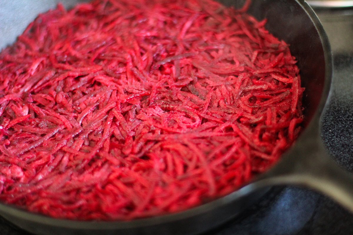 Cast iron skillet with beet hash browns inside, cooking on the stove top.