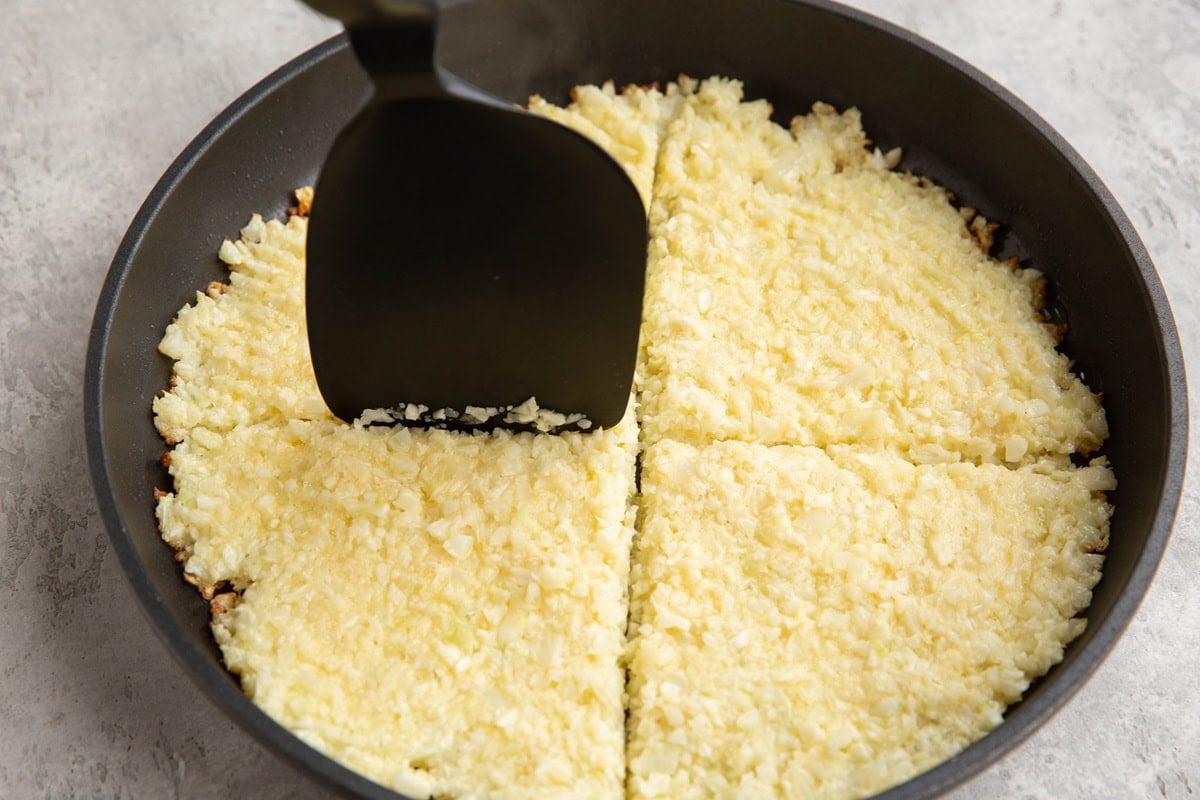 Spatula cutting the cauliflower hash browns into four sections to flip them.