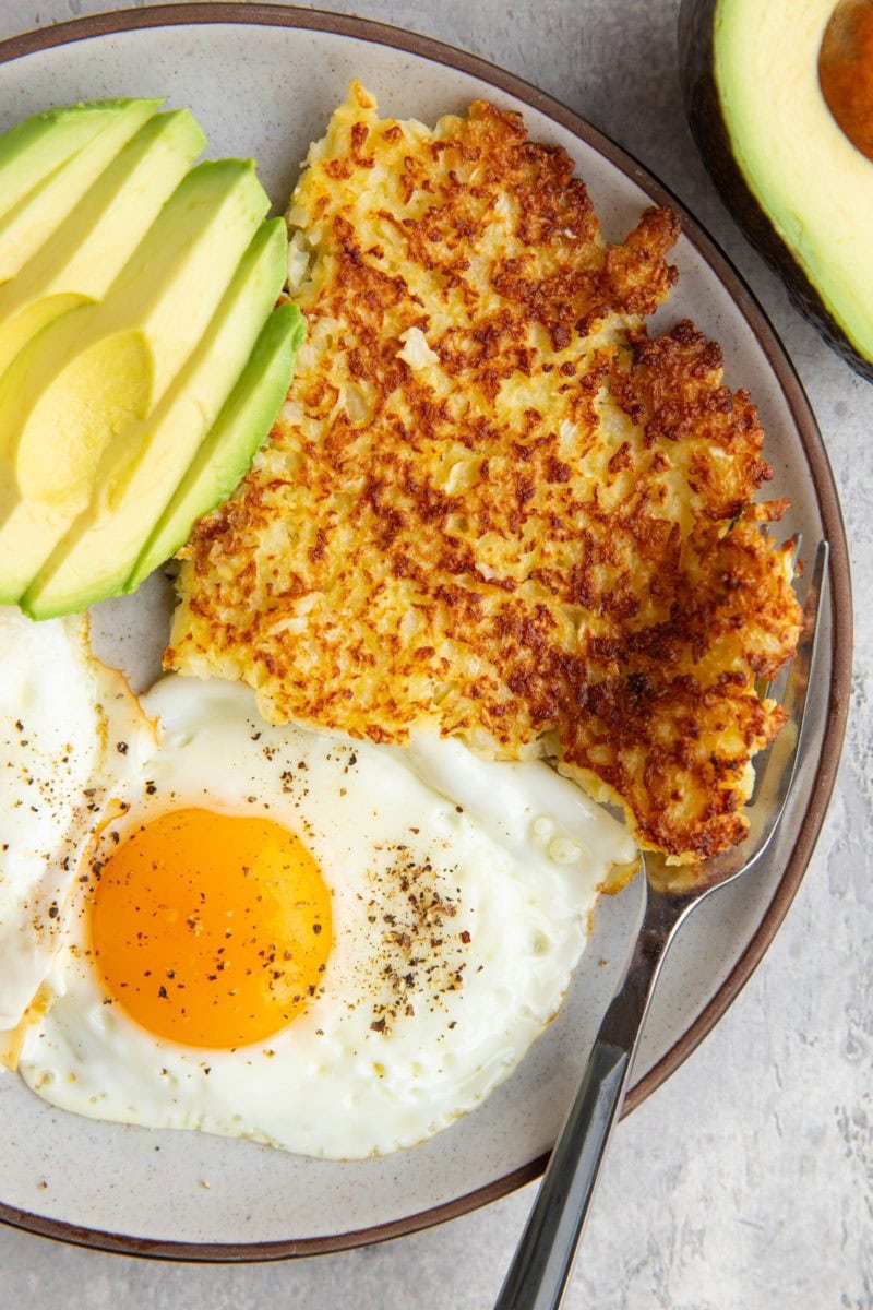 Cauliflower hash browns on a plate with sunny side up eggs and sliced avocado with a fork to the side.
