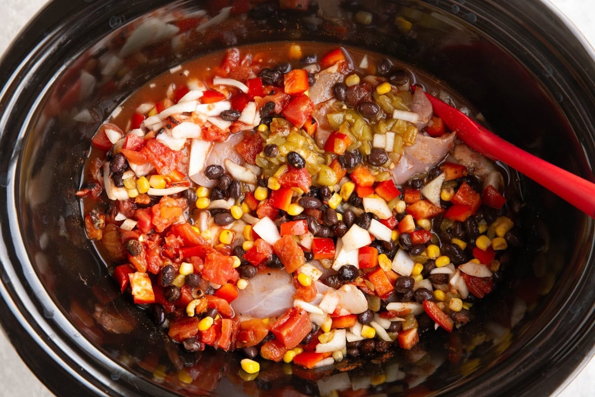 All of the ingredients for chicken chili stirred up inside of a slow cooker with a rubber spatula to the side.