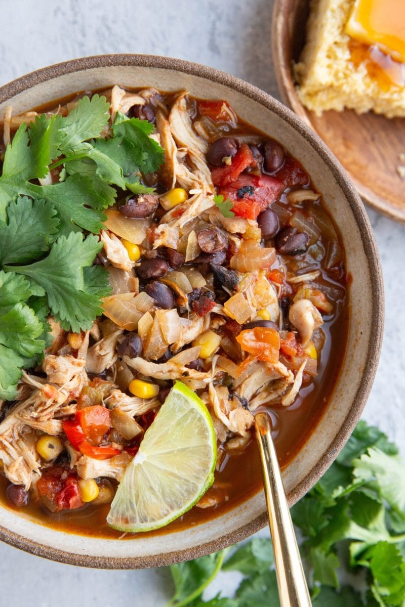 Large bowl full of shredded chicken chili with black beans and corn with fresh cilantro and a slice of lime on top. A plate of cornbread with butter and honey to the side.