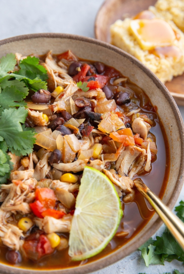 Big bowl full of shredded chicken chili with fresh cilantro and a slice of lime on top, ready to eat. A plate of corn bread with butter and honey in the background.