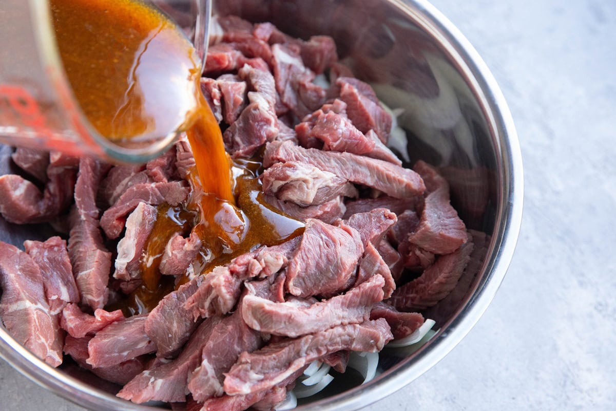 Stainless steel bowl full of strips of beef with shawarma marinade being poured into it.