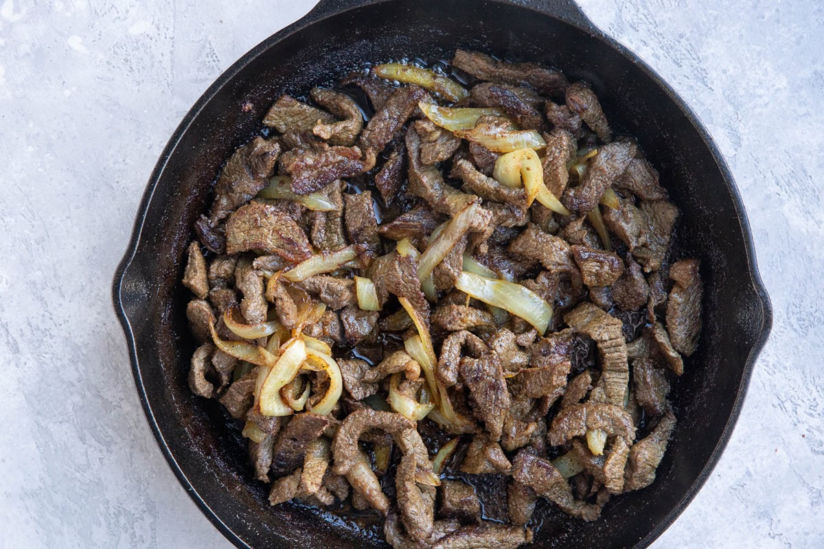 Cast iron skillet with strips of beef and onion in a cast iron skillet.