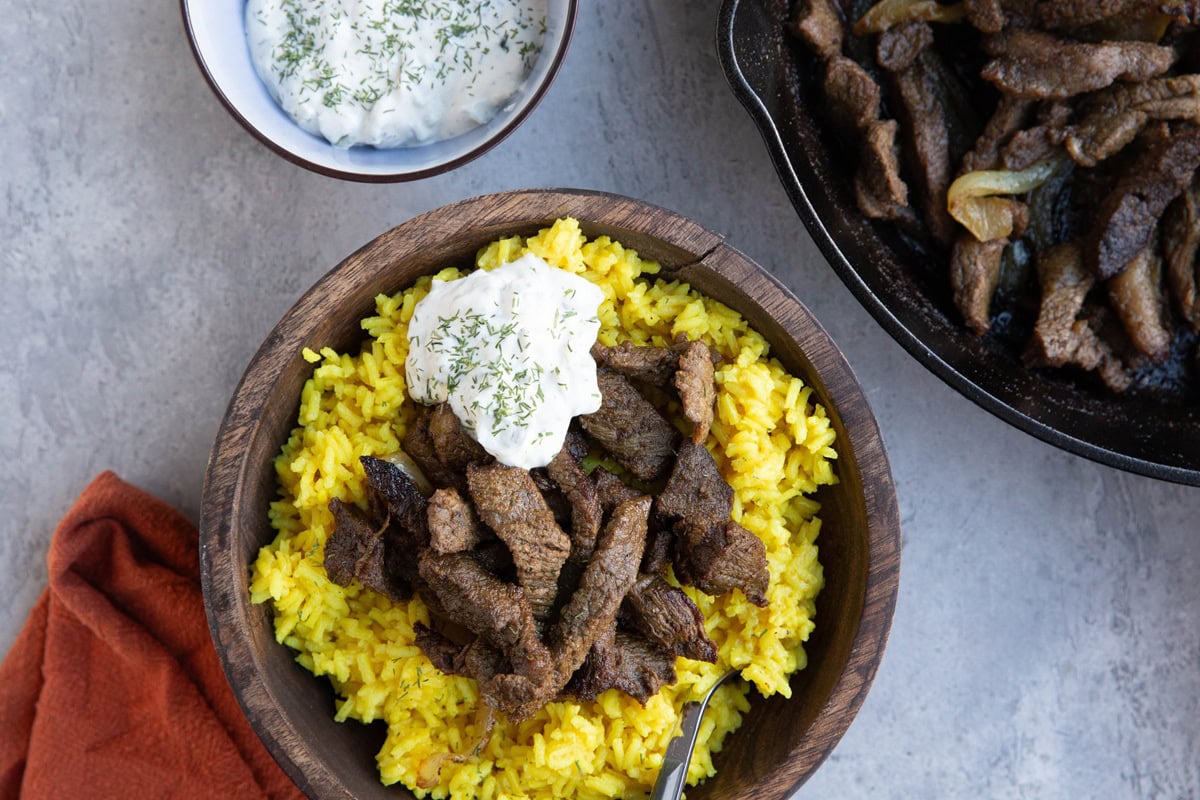 Wooden bowl full of yellow rice and beef shawarma with a skillet to the side that's full of more shawarma. A bowl of tzatziki sauce to the side.
