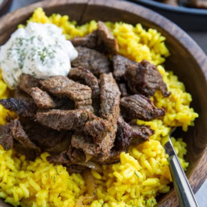 Large wooden bowl full of yellow rice and beef shawarma meat with tzatziki sauce on top and a wood inlay fork, ready to eat. The skillet with the rest of the meat is in the background.