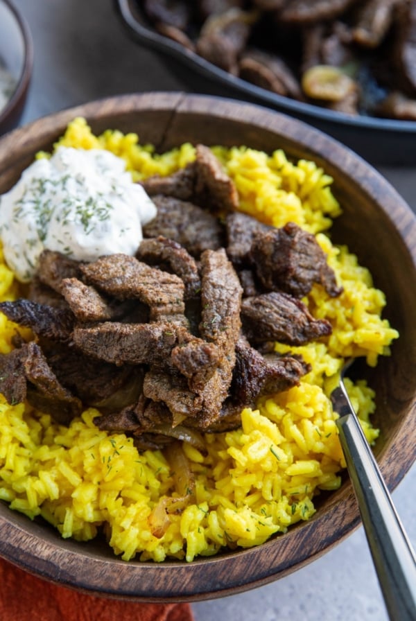 Large wooden bowl full of yellow rice and beef shawarma meat with tzatziki sauce on top and a wood inlay fork, ready to eat. The skillet with the rest of the meat is in the background.