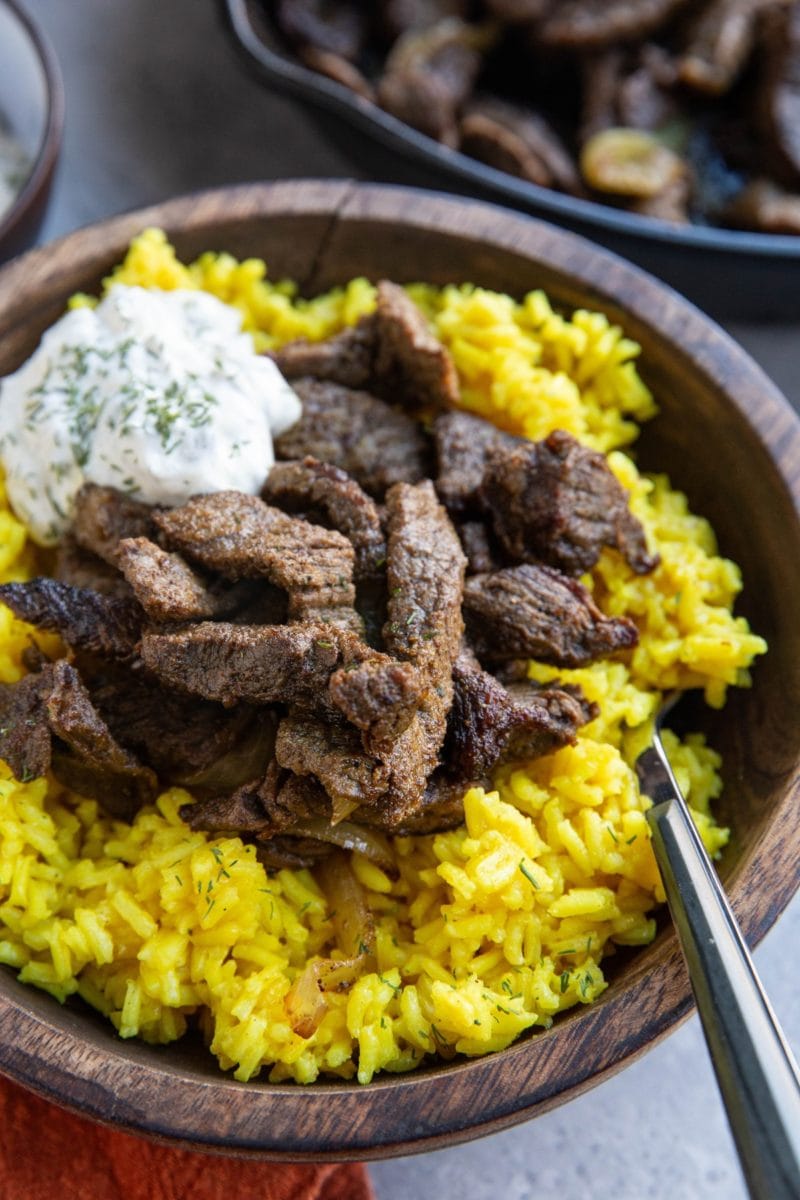 Large wooden bowl full of yellow rice and beef shawarma meat with tzatziki sauce on top and a wood inlay fork, ready to eat. The skillet with the rest of the meat is in the background.