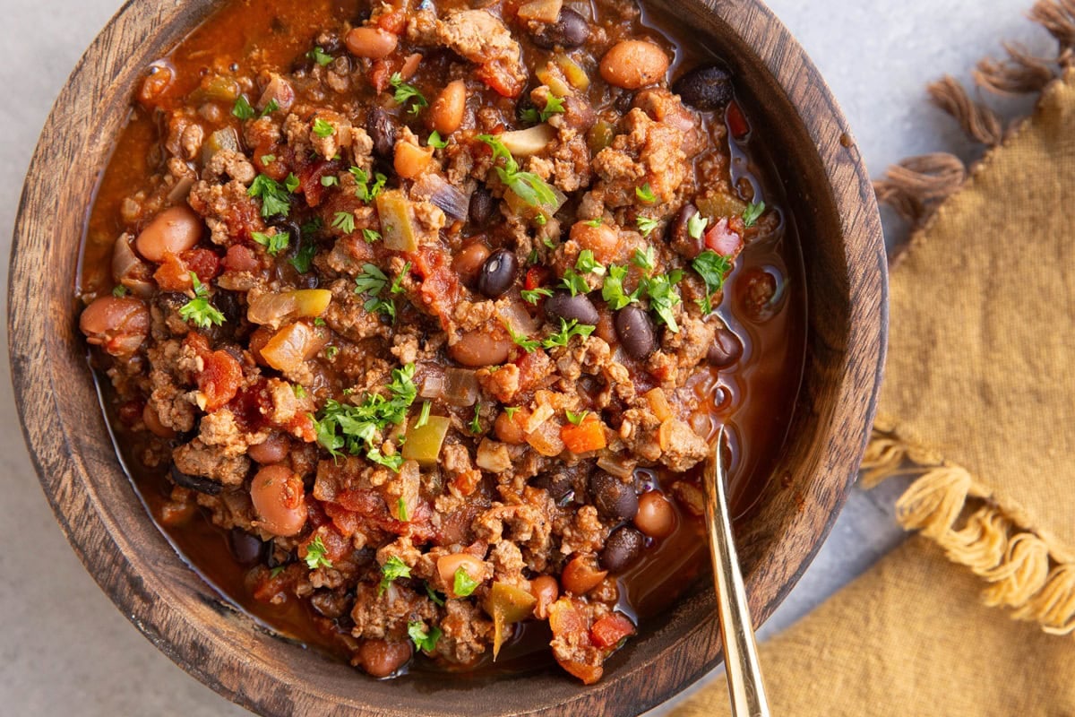 Elk chili in a large wooden bowl, sprinkled with chopped parsley. A golden spoon in the bowl and a golden napkin to the side, ready to eat.