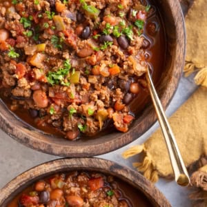 Two large wooden bowls full of elk chili with chopped parsley on top and a golden spoon and a gold napkin to the side.