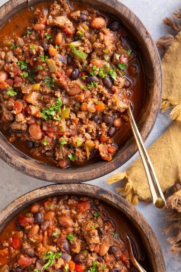 Two large wooden bowls full of elk chili with chopped parsley on top and a golden spoon and a gold napkin to the side.