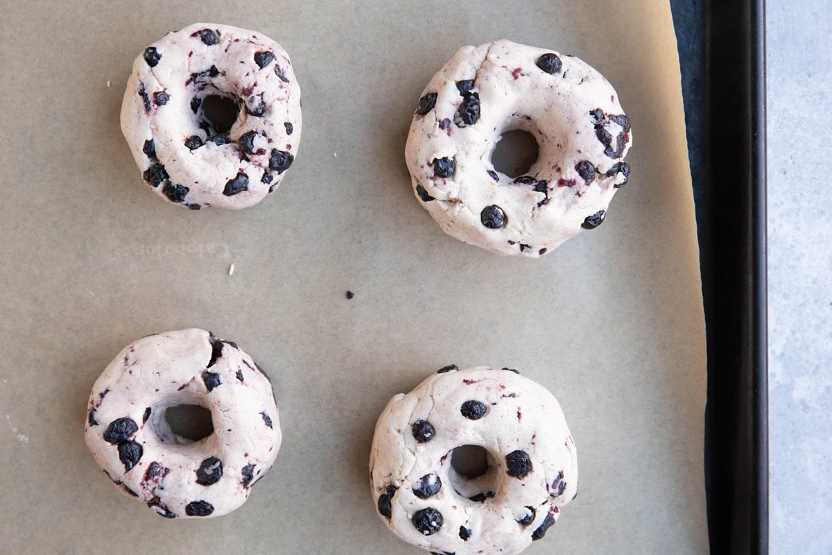 Four raw blueberry bagels on a parchment lined baking sheet, ready to bake the dough.