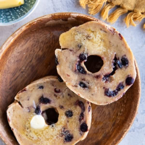 A blueberry bagel sliced in half covered with butter with a golden napkin to the side and a dish of butter.