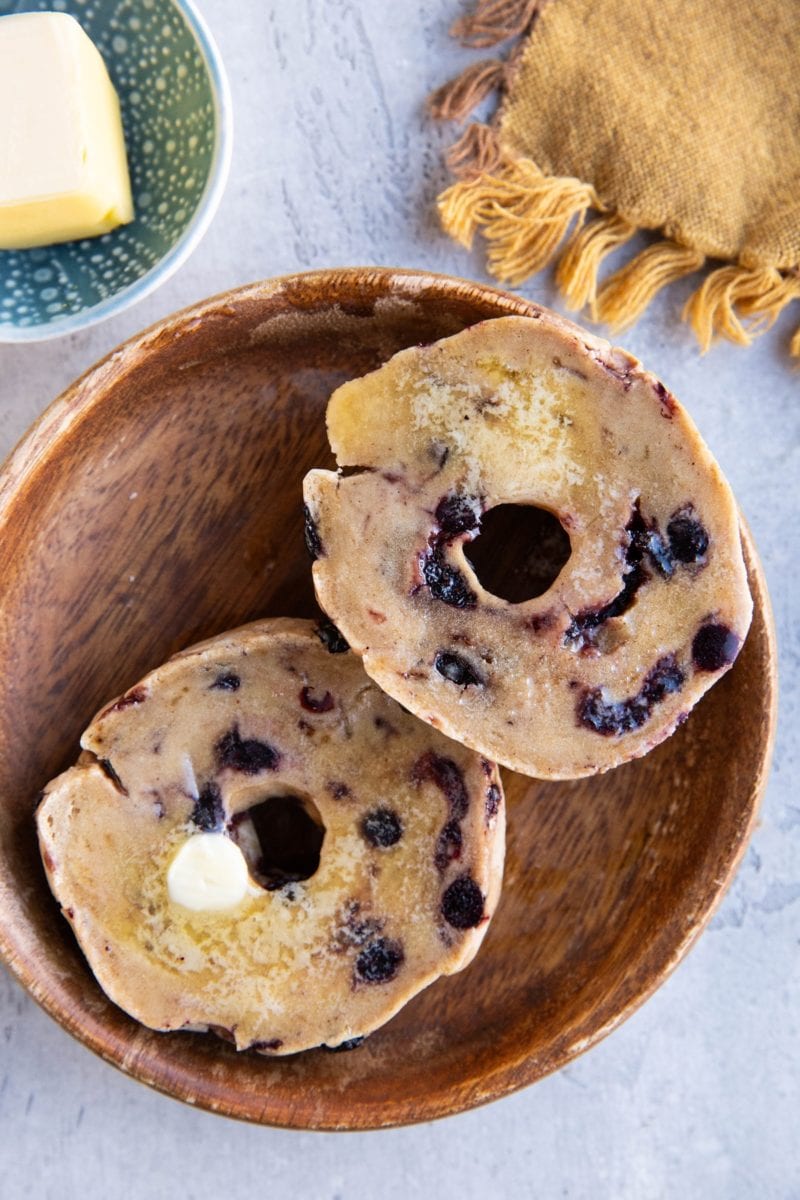 A blueberry bagel sliced in half covered with butter with a golden napkin to the side and a dish of butter.
