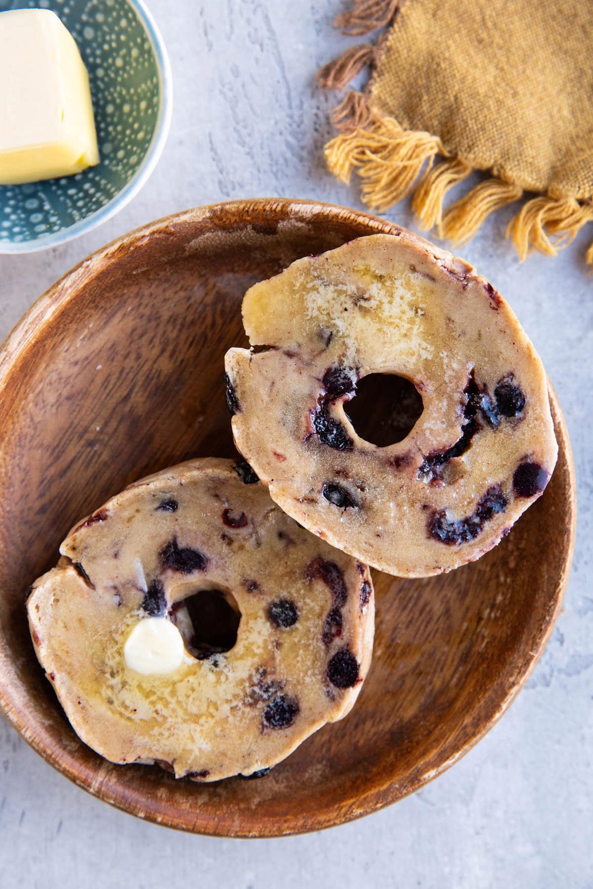 A blueberry bagel sliced in half covered with butter with a golden napkin to the side and a dish of butter.