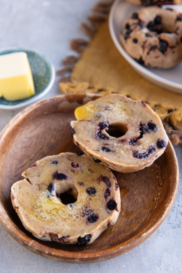 Wooden plate of a blueberry bagel sliced in half with melted butter on top. A plate of blueberry bagels in the background on top of a golden napkin.