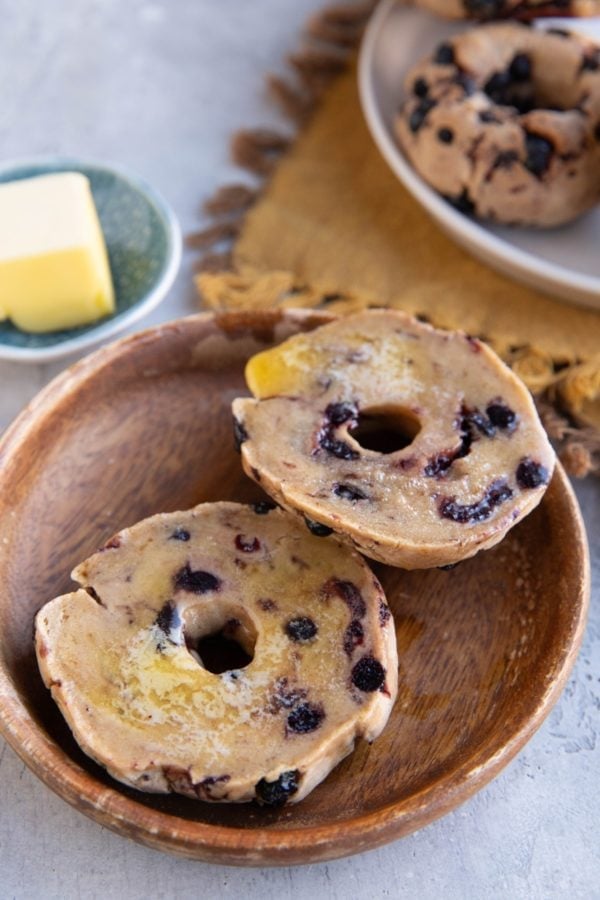 Wooden plate of a blueberry bagel sliced in half with melted butter on top. A plate of blueberry bagels in the background on top of a golden napkin.