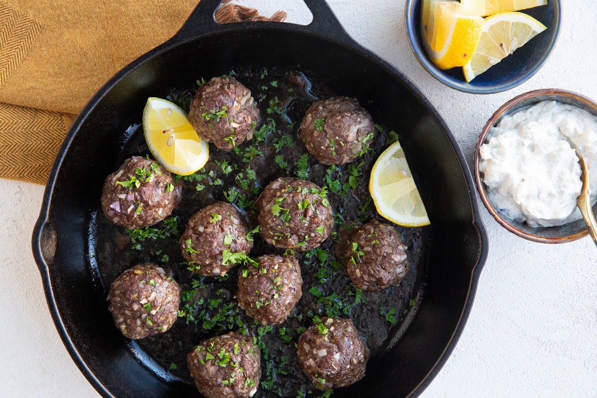 Cast iron skillet with meatballs sprinkled with chopped parsley and sliced lemons to the side. A bowl of more sliced lemons and a bowl of tzatziki sauce.