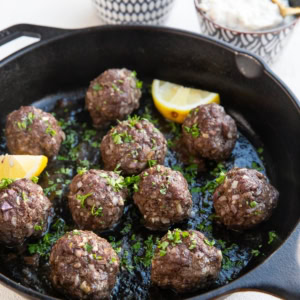 Cast iron skillet full of 10 meatballs that are sprinkled with fresh parsley. A bowl of sliced lemons and a bowl of tzatziki sauce in the background, ready to serve.