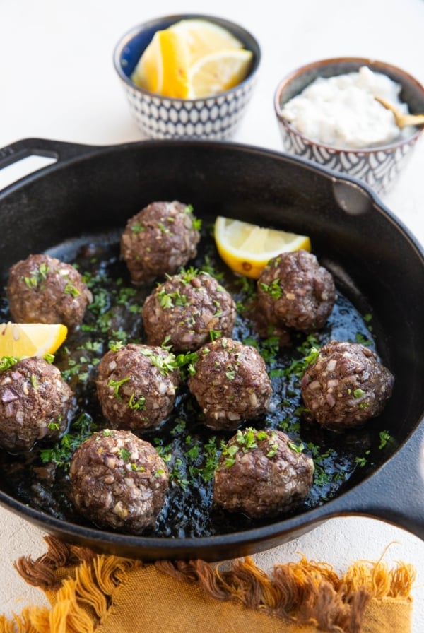 Cast iron skillet full of 10 meatballs that are sprinkled with fresh parsley. A bowl of sliced lemons and a bowl of tzatziki sauce in the background, ready to serve.