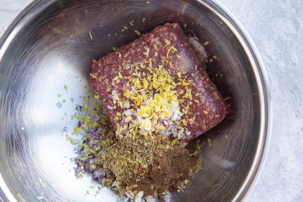 Meat, lemon zest, onion, garlic, and spices in a stainless steel mixing bowl, ready to mix together to make meatballs.