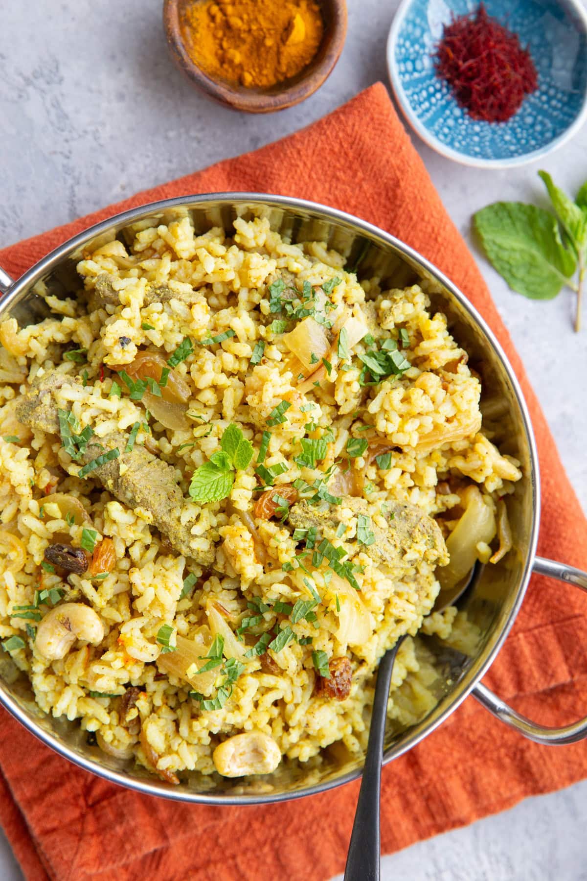 Stainless steel dish full of lamb biryani sprinkled with mint leaves. A dish of saffron threads and a small bowl of ground turmeric to the side.