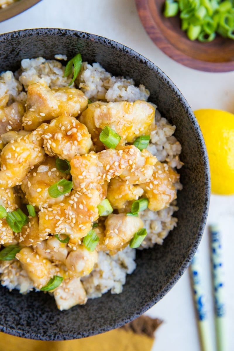 large bowl full of brown rice and lemon chicken with green onions and sesame seeds on top and chop sticks to the side. A wooden bowl sits to the side with chopped green onions.