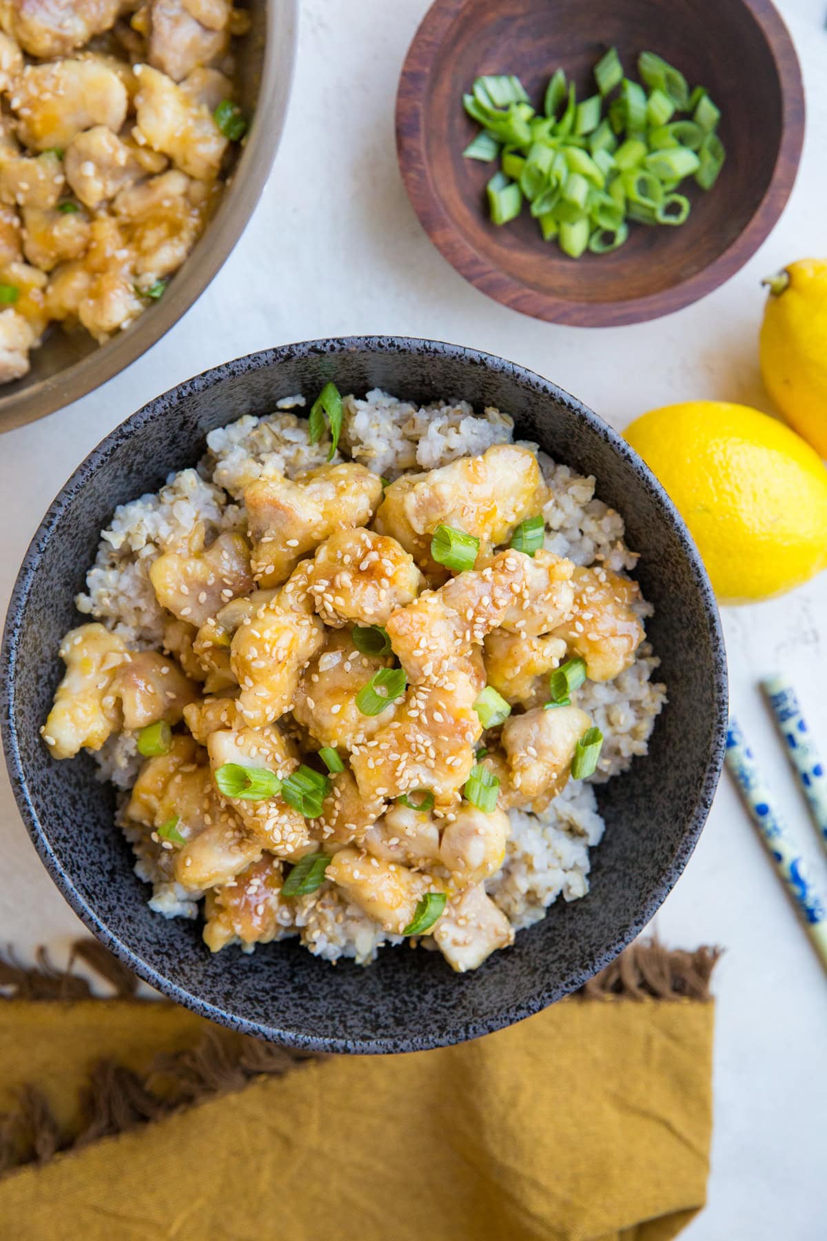 Large black speckled bowl full of lemon chicken on top of brown rice, sprinkled with green onions. The skillet with the rest of the chicken is off to the side, with fresh lemons, a bowl of green onions, and chop sticks.