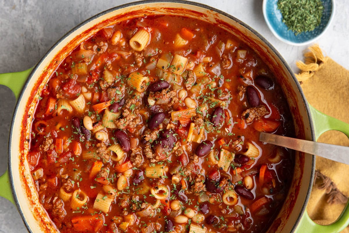 Large pot of pasta e fagioli soup with a ladle and a golden napkin to the side and a small blue bowl of dried parsley.