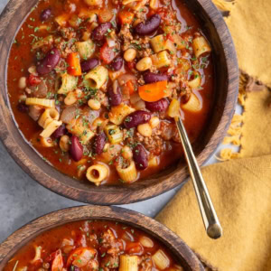 Two wooden bowls full of pasta e fagioli soup with golden spoons and a golden napkin to the side, ready to serve.