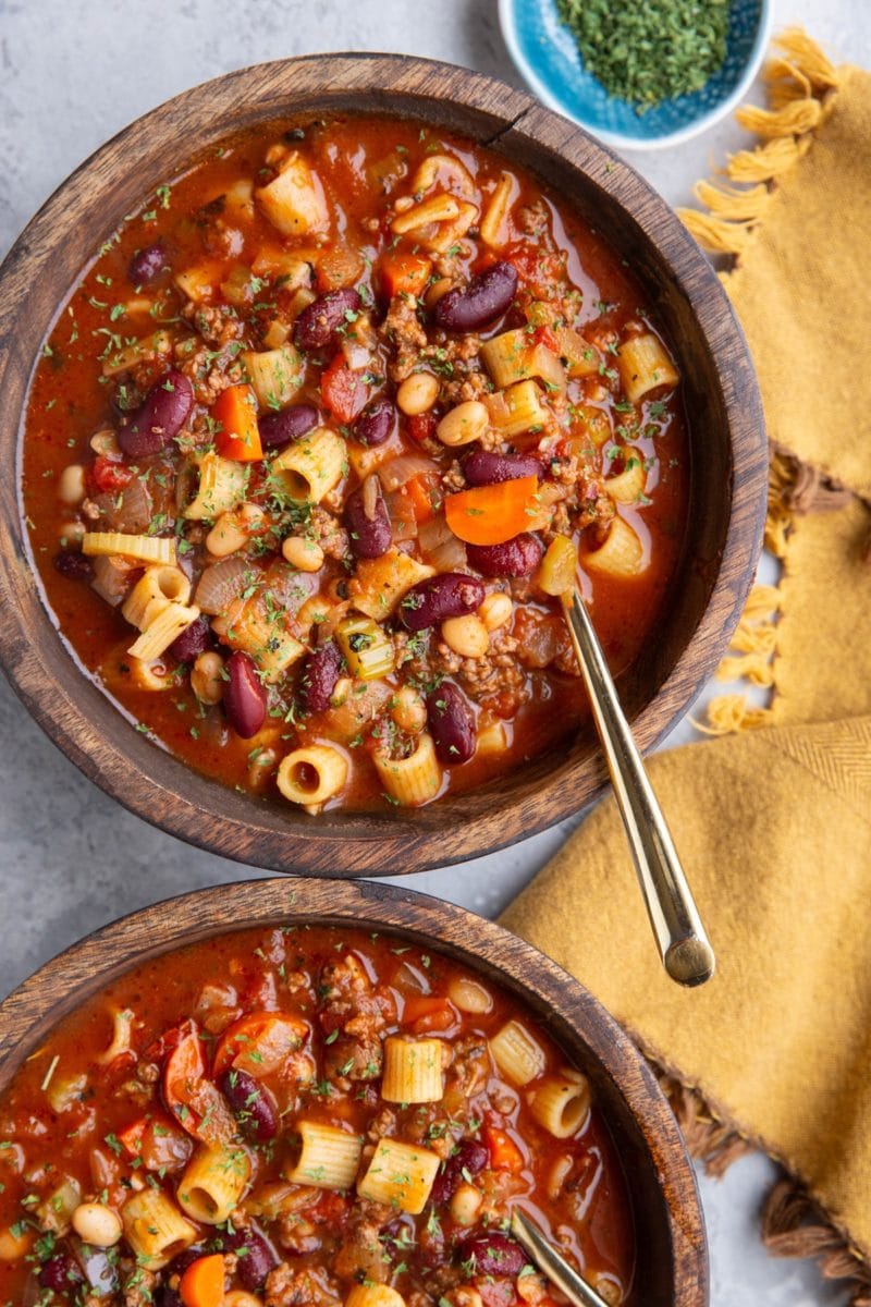Two wooden bowls full of pasta e fagioli soup with golden spoons and a golden napkin to the side, ready to serve.