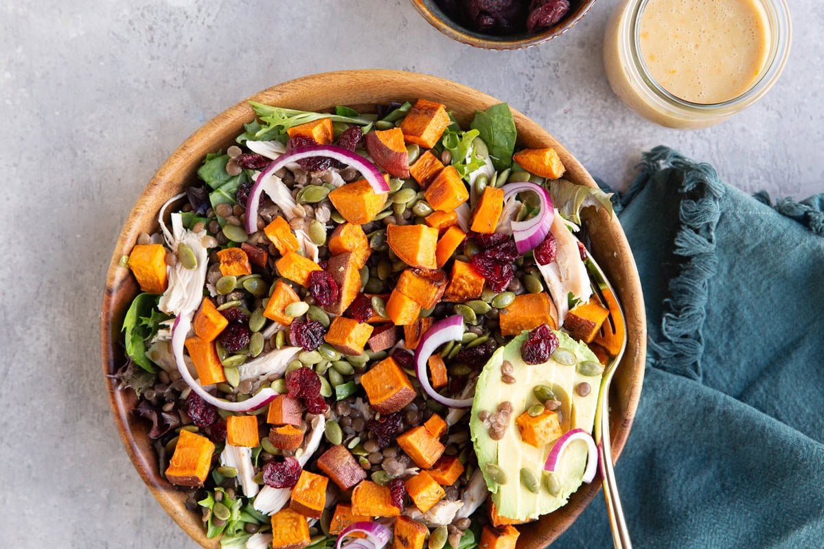 Wooden bowl full of a green salad with roasted sweet potatoes, chicken, avocado, pumpkin seeds, red onion, and dried cranberries. A jar of salad dressing to the side and a dark blue napkin, ready to serve.