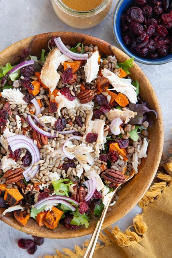 Large wooden bowl of salad with chicken, roasted sweet potatoes, lentils, pecans, dried cranberries, sunflower seeds, and red onions. a bowl of dried cranberries to the side and a jar of vinaigrette.