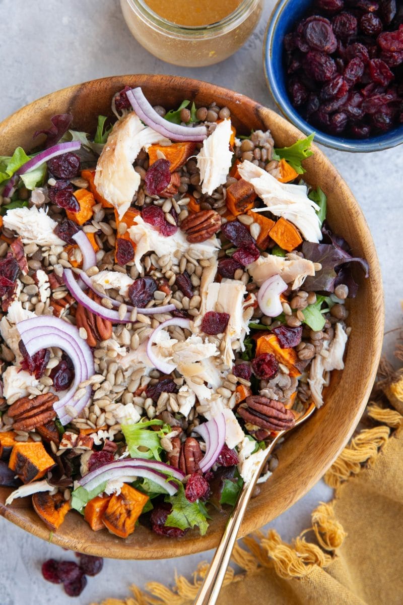 Large wooden bowl of salad with chicken, roasted sweet potatoes, lentils, pecans, dried cranberries, sunflower seeds, and red onions. a bowl of dried cranberries to the side and a jar of vinaigrette.