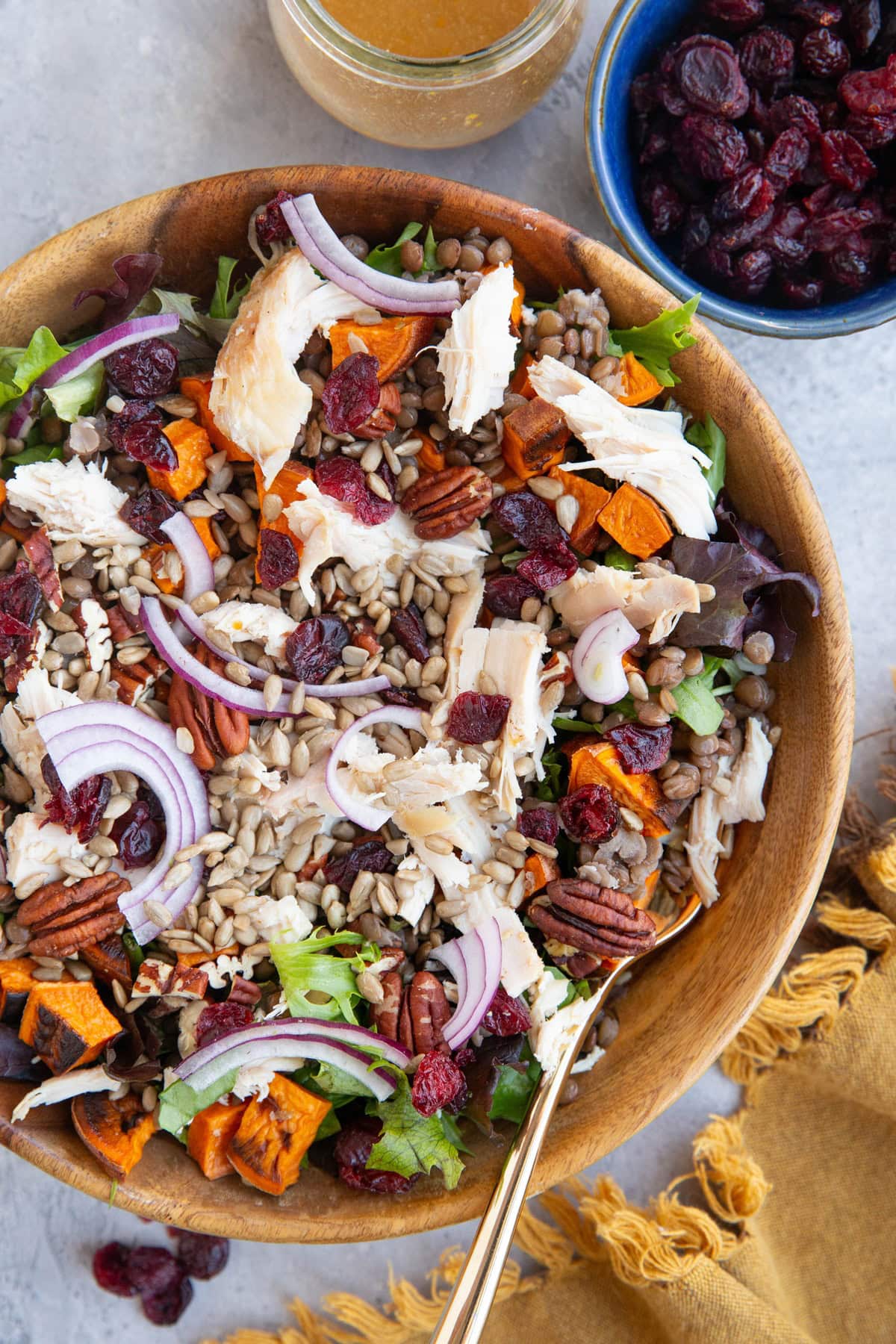 Large wooden bowl of salad with chicken, roasted sweet potatoes, lentils, pecans, dried cranberries, sunflower seeds, and red onions. a bowl of dried cranberries to the side and a jar of vinaigrette.