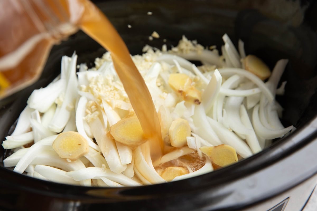 Pouring broth into the slow cooker with the onions, garlic, and ginger.