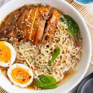 Big white bowl full of ramen noodles with broth, sliced chicken and soft boiled egg. Sprinkled with sesame seeds, red pepper flakes, and parsley with a black soup spoon to the side and a small blue bowl of sesame seeds.